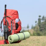 Red backpack with hiking pole and mat on grassy field with forest background.